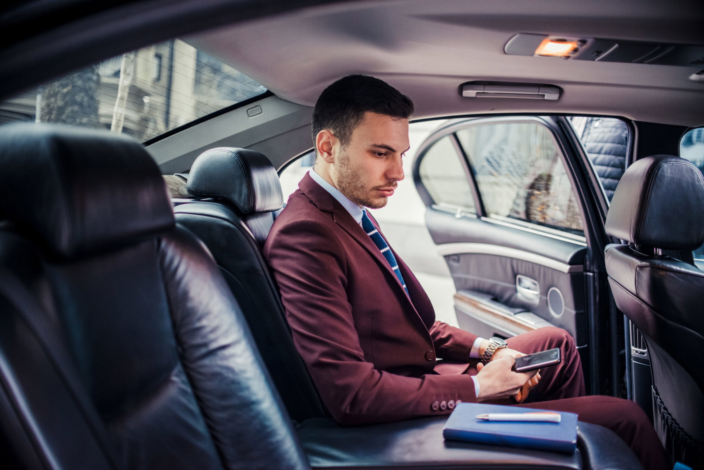A man in a maroon suit sits in the back seat of a limousine, looking at his phone, with a notebook and pen beside him. corporate transfer services