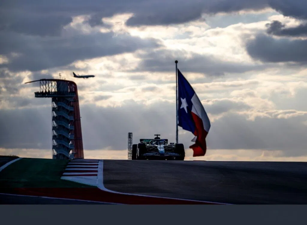 A Formula 1 car drives on a track at sunset during the Austin F1 race, passing a large Texas flag, with a plane and observation tower visible in the background.